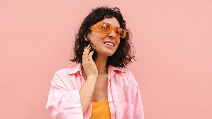 Young woman is smiling and posing against pink background. Brunette is wearing sunglasses, yellow top and pink shirt.